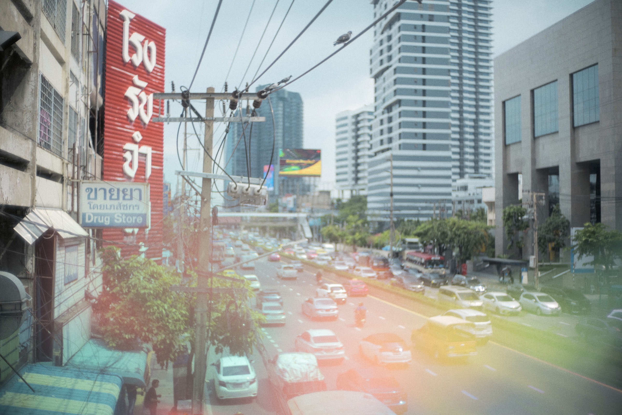 Bangkok traffic beside a red drug store sign with a soft rainbow light leak
